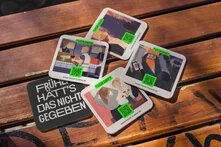 Foto von Holzbank, auf der Bierdeckel mit farbigen Illsutrationen und QR-Codes sind. Auf einem Bierdeckel steht in Weiß auf schwarzem Hintergrund: "Früher hätt`s das nicht gegeben"