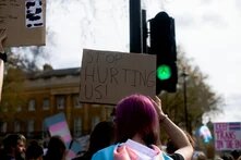 Personen mit langen lila Haaren hält ein Pappschild mit "Stop hurting us!" bei einer Demo hoch. Zu sehen sind Transgender-Pride-Flaggen.