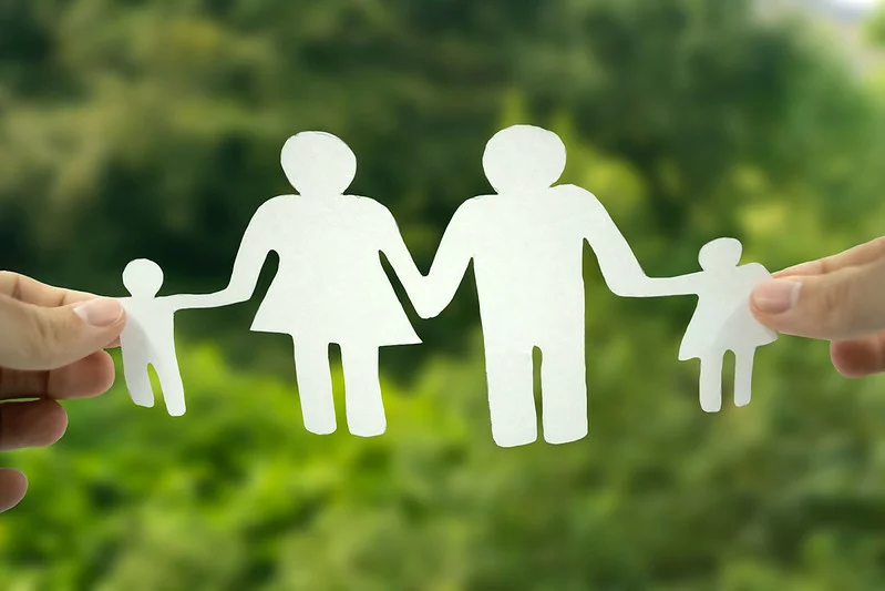 Grüner Hintergrund mit zwei Händen links und rechts, die aus weißem Papier ausgeschnittene Figuren halten, die auf eine hetero Kleinfamilie mit zwei Kindern hindeuten.